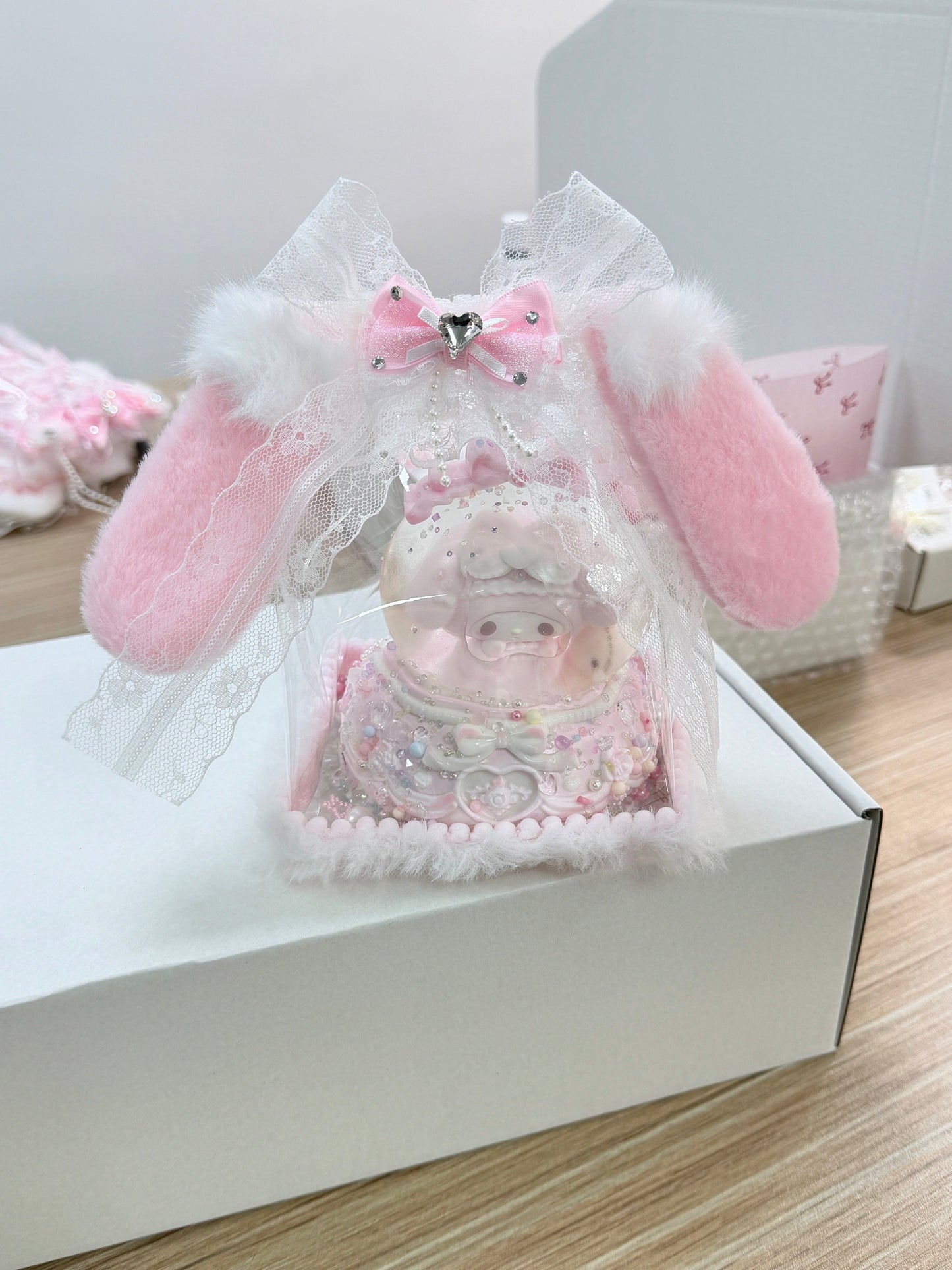 Luxury kawaii baby pig squeeze toy in a bunny ear shaped gift display with two large pink fluffy ears, white lace bow, heart crystal rhinestone, and clear acetate dome, sitting on a white gift box