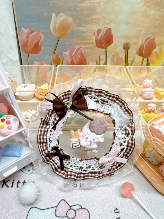 Kawaii baker lamb squeeze toy with whipped cream silicone texture, lilac clover crown, and baguette prop, displayed in a brown gingham and white lace wreath-style gift packaging with chocolate bow